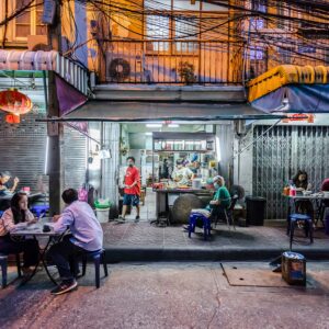 People dine at tables on a sidewalk outside a small street food restaurant in an urban area at night; the kitchen and staff are visible inside.
